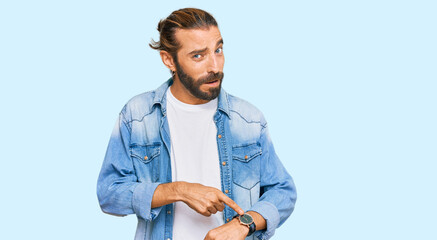 Attractive man with long hair and beard wearing casual denim jacket in hurry pointing to watch...