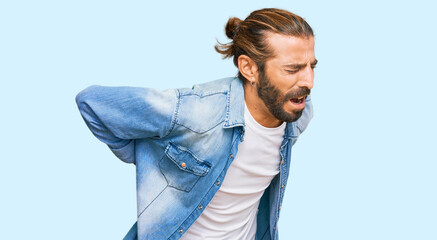 Attractive man with long hair and beard wearing casual denim jacket suffering of backache, touching back with hand, muscular pain