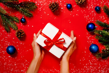 Woman hands hold a gift box decorated with tree branches, pine cones, snow and balls on a red...