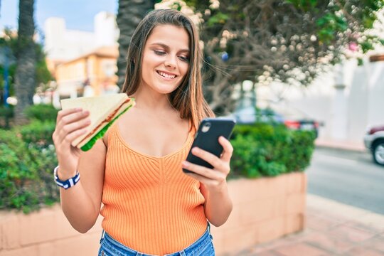 Young middle east girl smiling happy using smartphone and eating sandwich at the city.