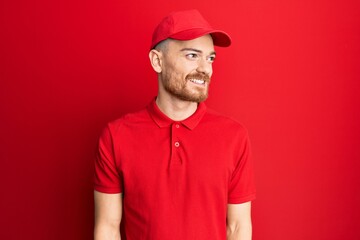 Young redhead man wearing delivery uniform and cap looking away to side with smile on face, natural expression. laughing confident.