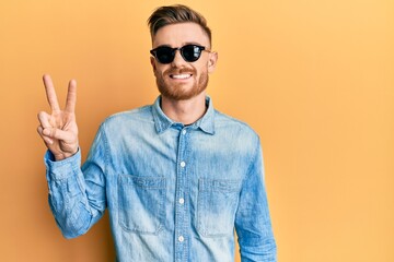 Naklejka premium Young redhead man wearing stylish sunglasses smiling with happy face winking at the camera doing victory sign. number two.