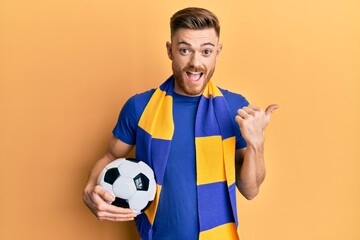 Young redhead man football hooligan holding ball pointing thumb up to the side smiling happy with open mouth