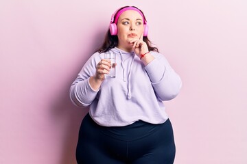 Young plus size woman drinking glass of water using headphones serious face thinking about question with hand on chin, thoughtful about confusing idea