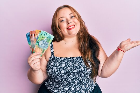 Plus size caucasian young woman holding australian dollars celebrating achievement with happy smile and winner expression with raised hand