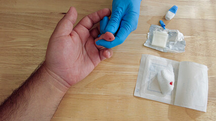 A medical personnel wearing gloves is performing a finger prick blood draw before a diagnostic testing at a doctors office. Used lancet and blood contaminated gauze pad is on the table