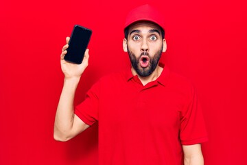 Young handsome man with beard wearing delivery uniform holding smartphone scared and amazed with open mouth for surprise, disbelief face