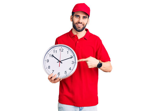 Young handsome man with beard wearing delivery uniform holding big clock smiling happy pointing with hand and finger