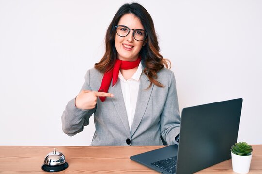 Young beautiful brunette woman working at hotel reception using laptop pointing finger to one self smiling happy and proud
