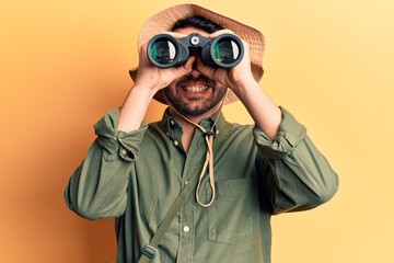 Young hispanic man wearing explorer hat holding binoculars looking positive and happy standing and...
