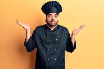Young hispanic man wearing cooker uniform clueless and confused with open arms, no idea and...