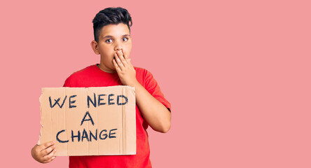 Little boy kid holding we need a change banner covering mouth with hand, shocked and afraid for...