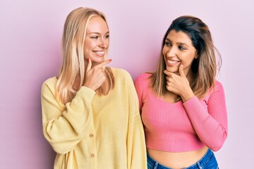 Two friends standing together over pink background with hand on chin thinking about question, pensive expression. smiling with thoughtful face. doubt concept.