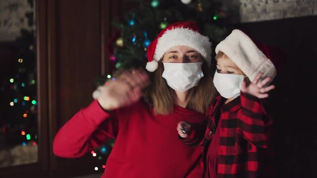 coronavirus, covid-19 in the new year. little daughter with her mother in a bad Santa hat and medical mask waving their hands at home under the Christmas tree.