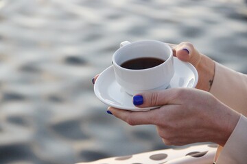 White cup of tea with a saucer in the graceful hands of a woman with a classic blue manicure and in beige clothes