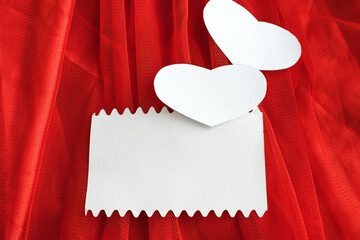 A white rectangular leaf and two white hearts are placed on a red background of tulle and satin material. Holiday background with copy space.