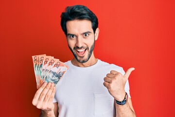 Young hispanic man holding australian dollars pointing thumb up to the side smiling happy with open mouth