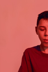 Obraz premium Cropped portrait of teenaged disabled boy with cerebral palsy keeping his eyes closed, looking thoughtful while posing isolated over red light background