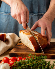 Faceless man cutting fresh home baked crusty bread with large knife on wooden Board on kitchen table. Homemade bakery, vertical