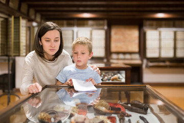 Fototapeta na wymiar Young female tutor with boy looking at exposition in glass case in historical museum