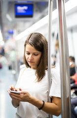 Portrait of positive girl standing in underground coach and using mobile phone