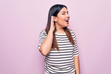 Young beautiful brunette woman wearing casual striped t-shirt over isolated pink background smiling with hand over ear listening and hearing to rumor or gossip. Deafness concept.