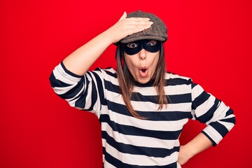 Young beautiful brunette burglar woman wearing cap and mask over isolated red background surprised with hand on head for mistake, remember error. Forgot, bad memory concept.