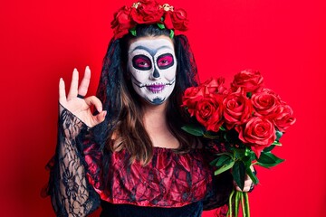Young woman wearing day of the dead costume holding flowers doing ok sign with fingers, smiling friendly gesturing excellent symbol