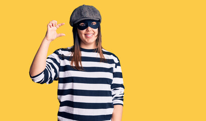 Young beautiful brunette woman wearing burglar mask smiling and confident gesturing with hand doing small size sign with fingers looking and the camera. measure concept.
