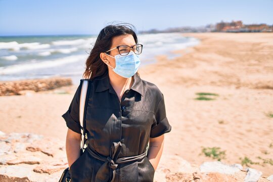 Young beautiful brunette woman wearing medical mask and glasses walking at the beach