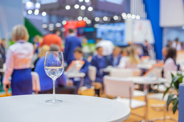 Close up view: glass of water, champagne or wine on white table against blurred business meeting, conference, seminar background. Corporate and communication concept