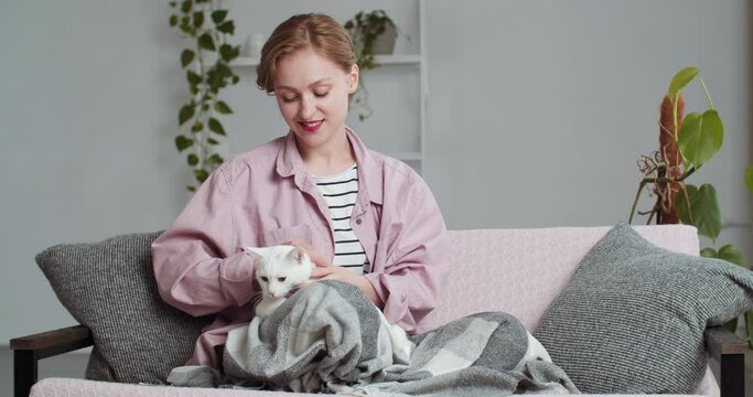 Blonde woman in pink casual shirt hiding behind warm sixth blanket sits on sofa in living room at home hugs stroking soft fluffy white cat animal pet kitten spends time calmly resting relaxing alone