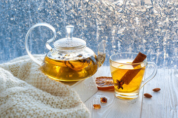 Winter warming tea with lemon and orange against the background of frosty patterns. Hot drink in a glass kettle and a cup on a wooden table in the background of the window.