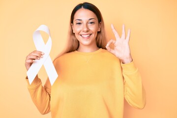 Young brunette woman holding cancer awareness white ribbon doing ok sign with fingers, smiling friendly gesturing excellent symbol