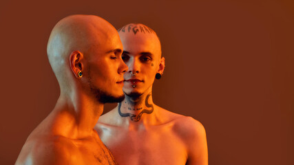 Fototapeta premium Front and side view of young half naked twin brothers with tattoos and piercings posing together, standing isolated over orange background