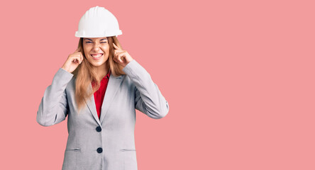 Beautiful young woman wearing architect hardhat covering ears with fingers with annoyed expression for the noise of loud music. deaf concept.