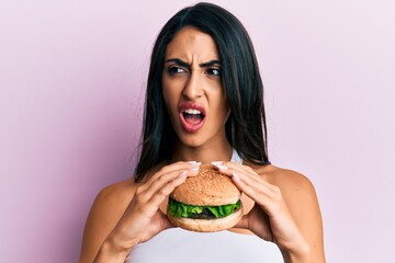 Beautiful hispanic woman eating a tasty classic burger angry and mad screaming frustrated and...