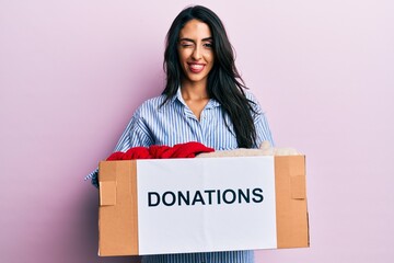 Beautiful hispanic woman volunteer holding donations box winking looking at the camera with sexy...