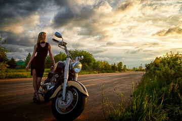 Attractive female brunette motorcyclist with motorcycle in a summer evening during sunset. Beautiful girl on nature and sky with sun and clouds background. Adventure concept