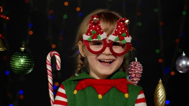 Child in glasses dancing, fooling around, making funny faces. Little blonde kid teen teenager girl in Christmas elf Santa helper costume posing on black background. New Year holidays celebration