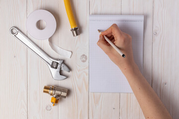 Female plumber preparing for work. Female hand with a pen, notebook, gas tap, gas hose and accessories for connection. On white wooden background.
