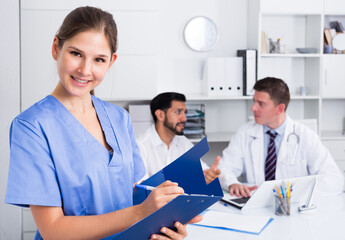 Smiling attractive female doctor writing notes on clipboard in office with colleagues