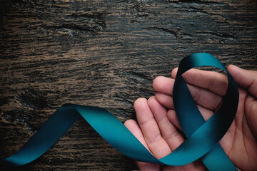Top view of hand holding teal ribbon on dark background with copy space. Cervical and ovarian cancer, sexual assault, pcos, ptsd, anxiety disorder, agoraphobia and scleroderma awareness concept.