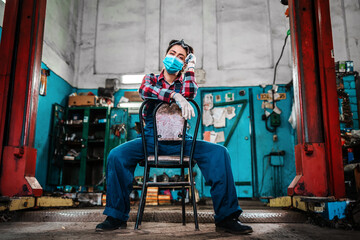 A young female mechanic in uniform, wearing gloves and a medical mask, poses sitting on a chair.In the background there is an auto repair shop and a lift for cars