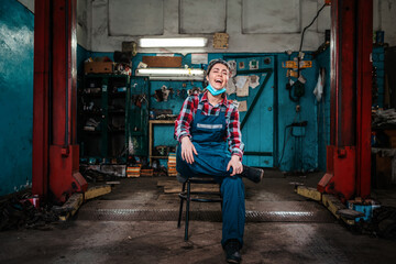Fototapeta na wymiar A young female mechanic in a work uniform sits on a chair with a medical mask on her chin and laughs. Indoor. Garage