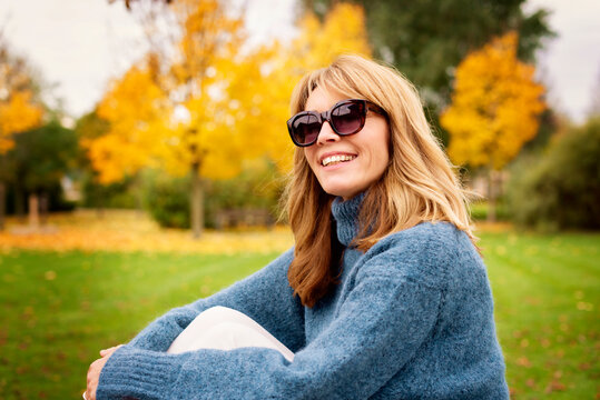 Portrait shot of happy mature woman wearing sunglasses and turtleneck sweater while relaxing in the park at autumn.