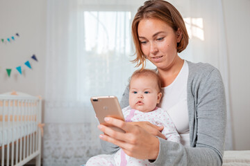 Portrait of young mother holds a cute baby in her arms and shows him something on her smartphone....