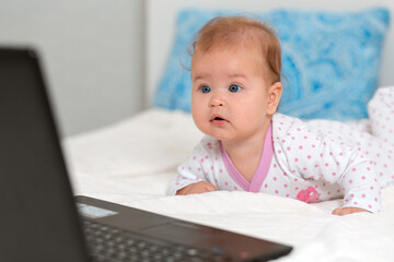 Portrait of amazement baby in sliders lies on the bed and looks at the laptop. The concept of teaching children to modern technologies