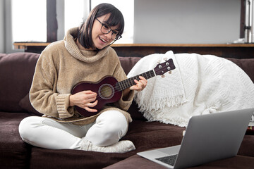 Young beautiful woman learning to play ukulele at home with online lessons. - obrazy, fototapety, plakaty