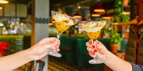 Two young women holding glasses with dessert over blurred restaurant background, selective focus.